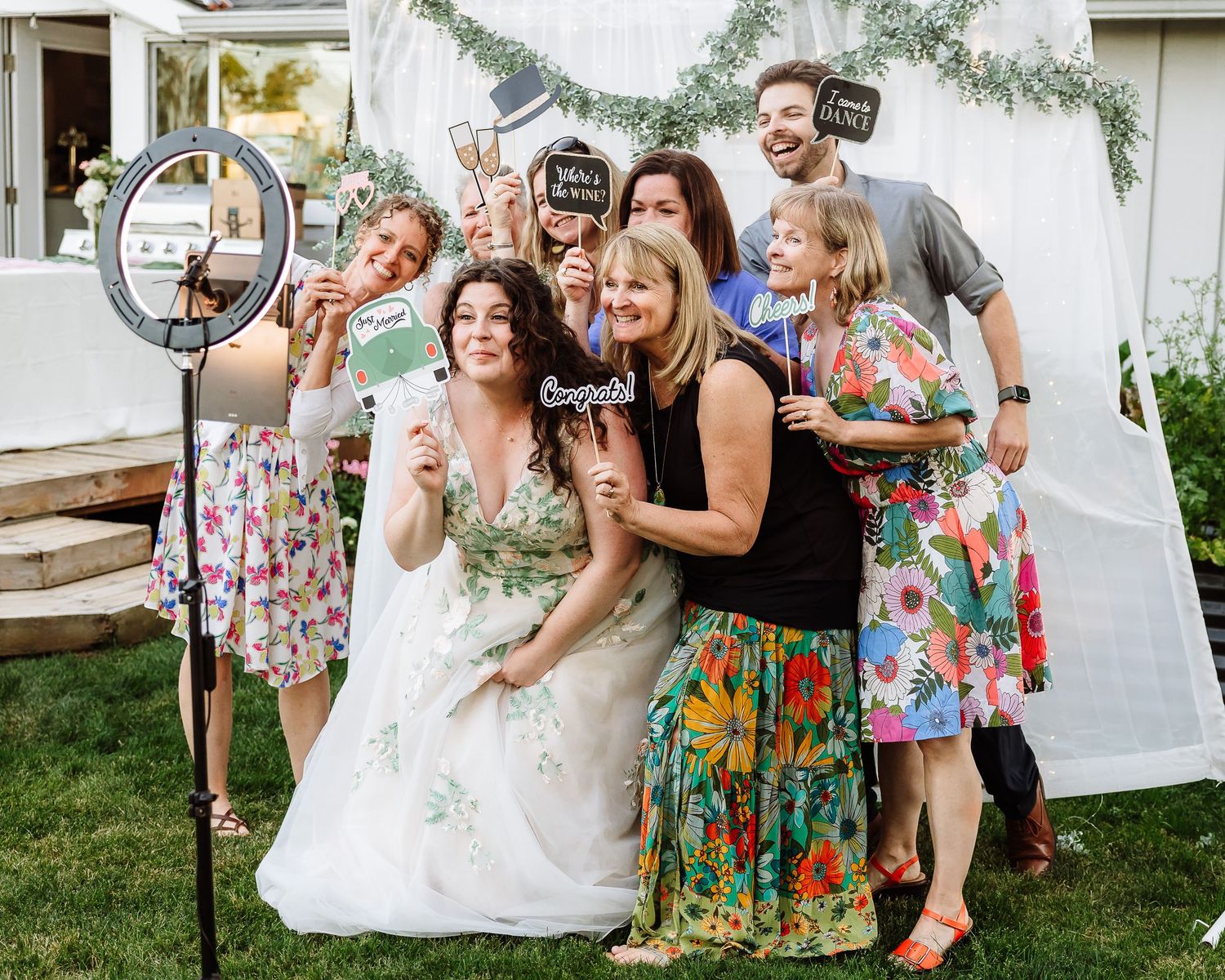 Bride poses with wedding guests at a photo booth with ring light and fun props at outdoor reception.