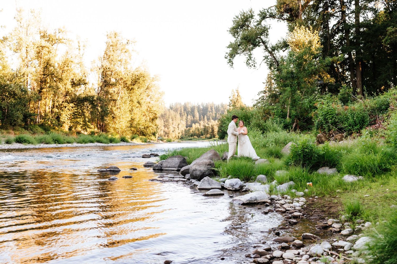 Newlyweds sharing a romantic kiss on a rocky riverbank surrounded by golden autumn trees at sunset.