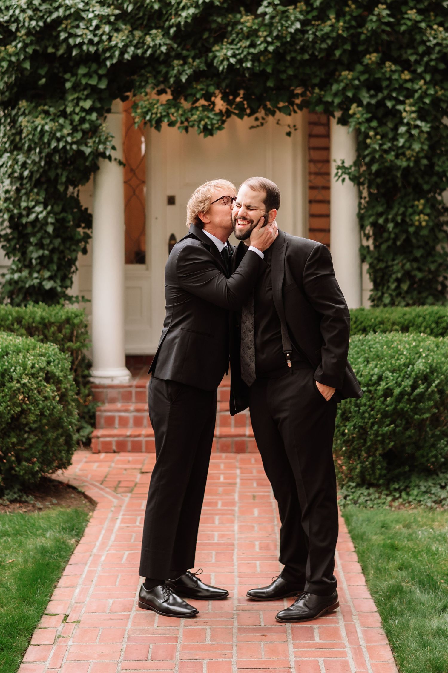 Two men in black suits share a tender moment on a brick pathway surrounded by lush greenery.