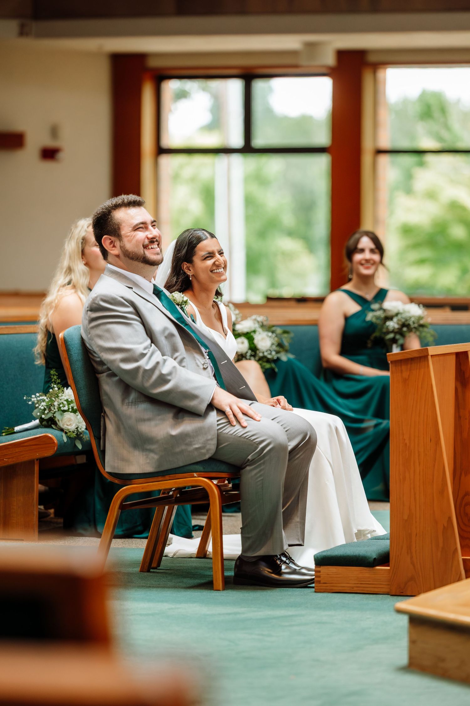 Smiling bride and groom sit together at altar during church wedding ceremony with bridesmaids behind.