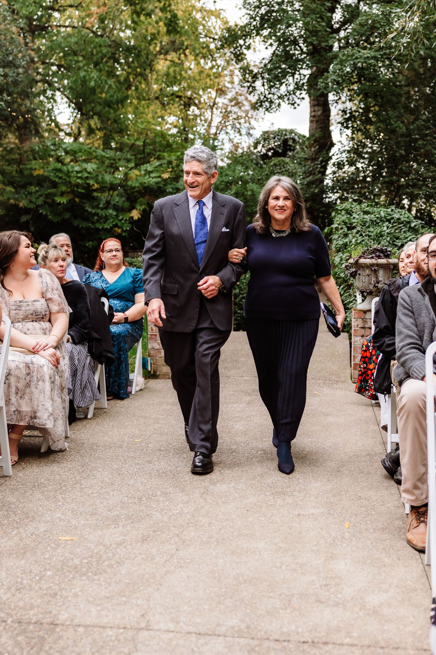 Elegant couple walks down outdoor wedding aisle surrounded by seated guests and lush green trees.