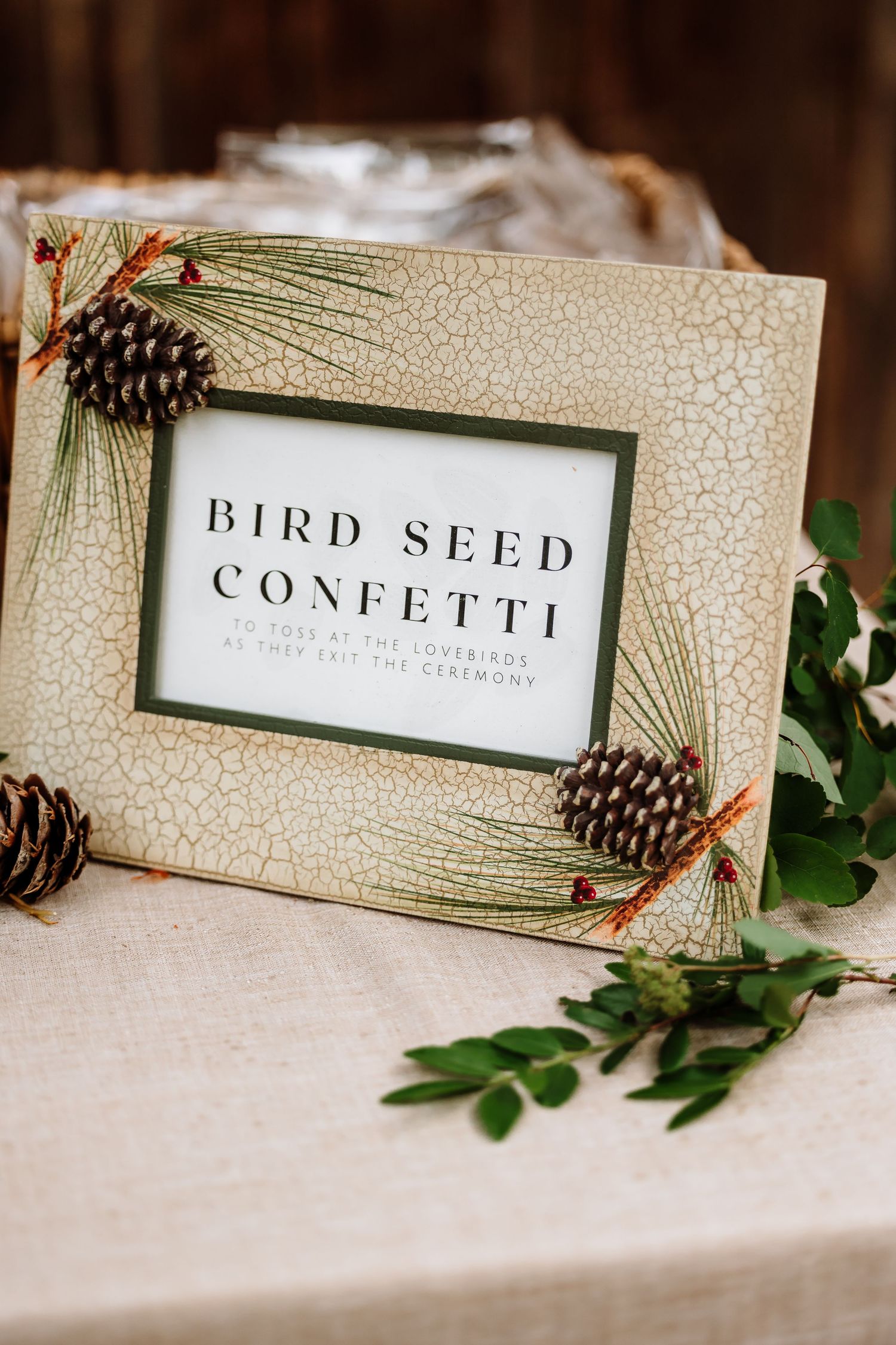 Bird seed confetti sign in pine cone decorated frame on linen table at rustic wedding.