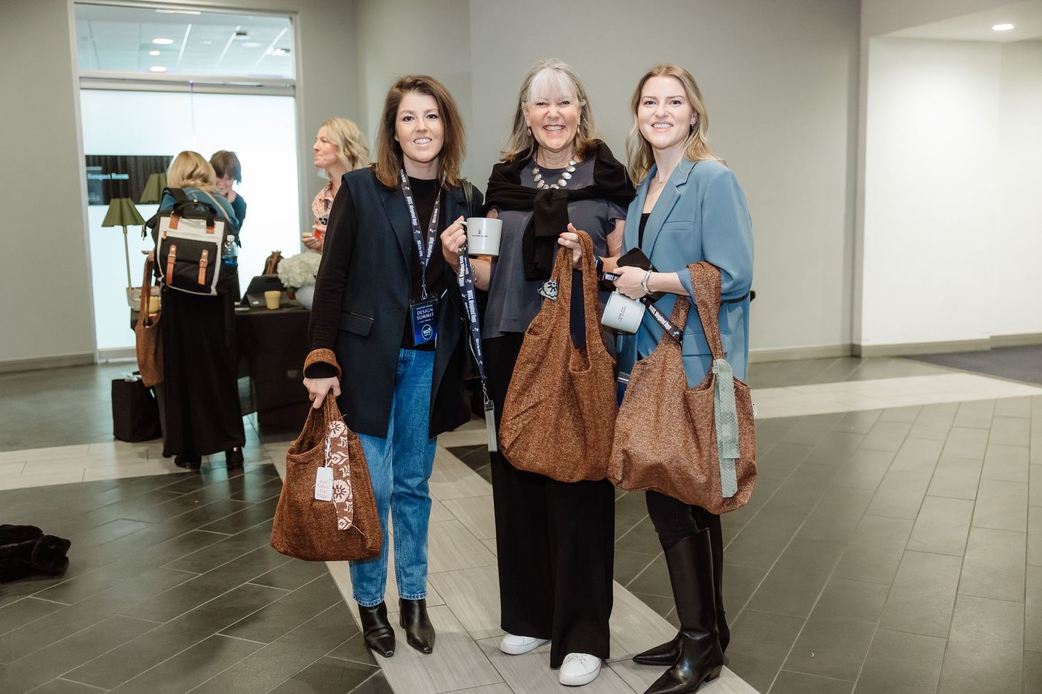 Three women smiling and holding brown tote bags at an indoor conference event.