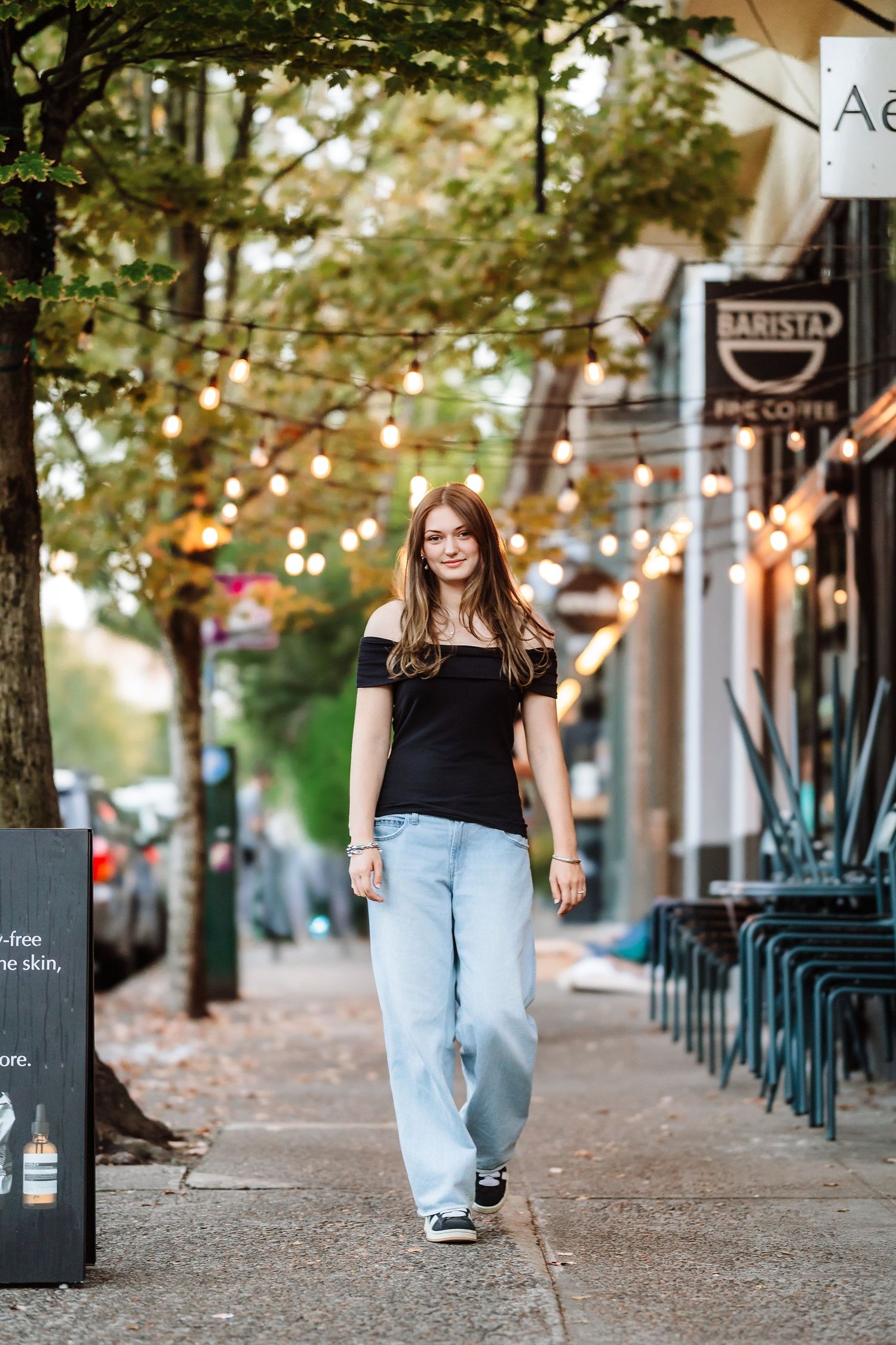 Teen girl in black off-shoulder top and wide-leg jeans walking on a city sidewalk.