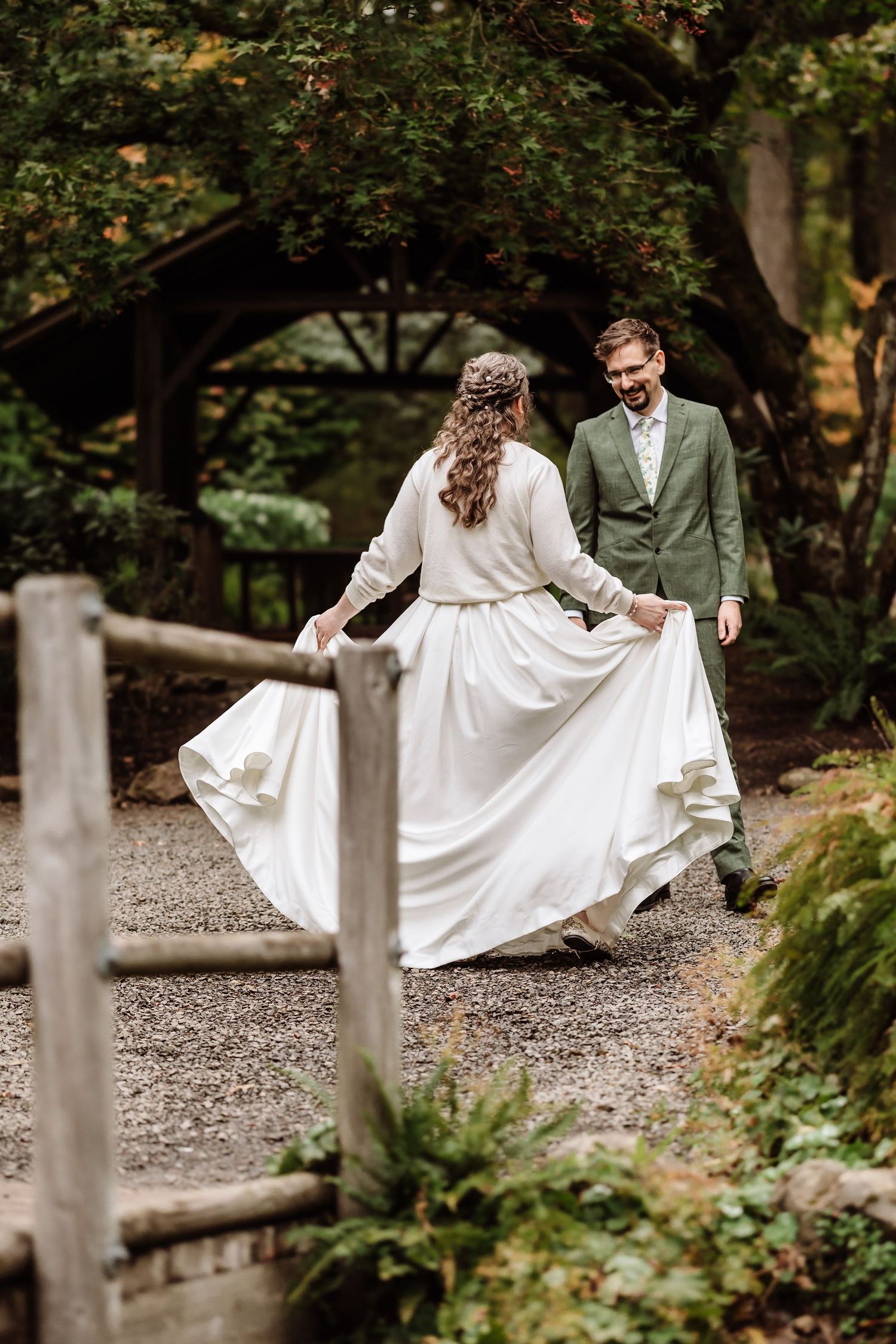 Bride twirls white gown while groom in green suit smiles on garden gravel path.