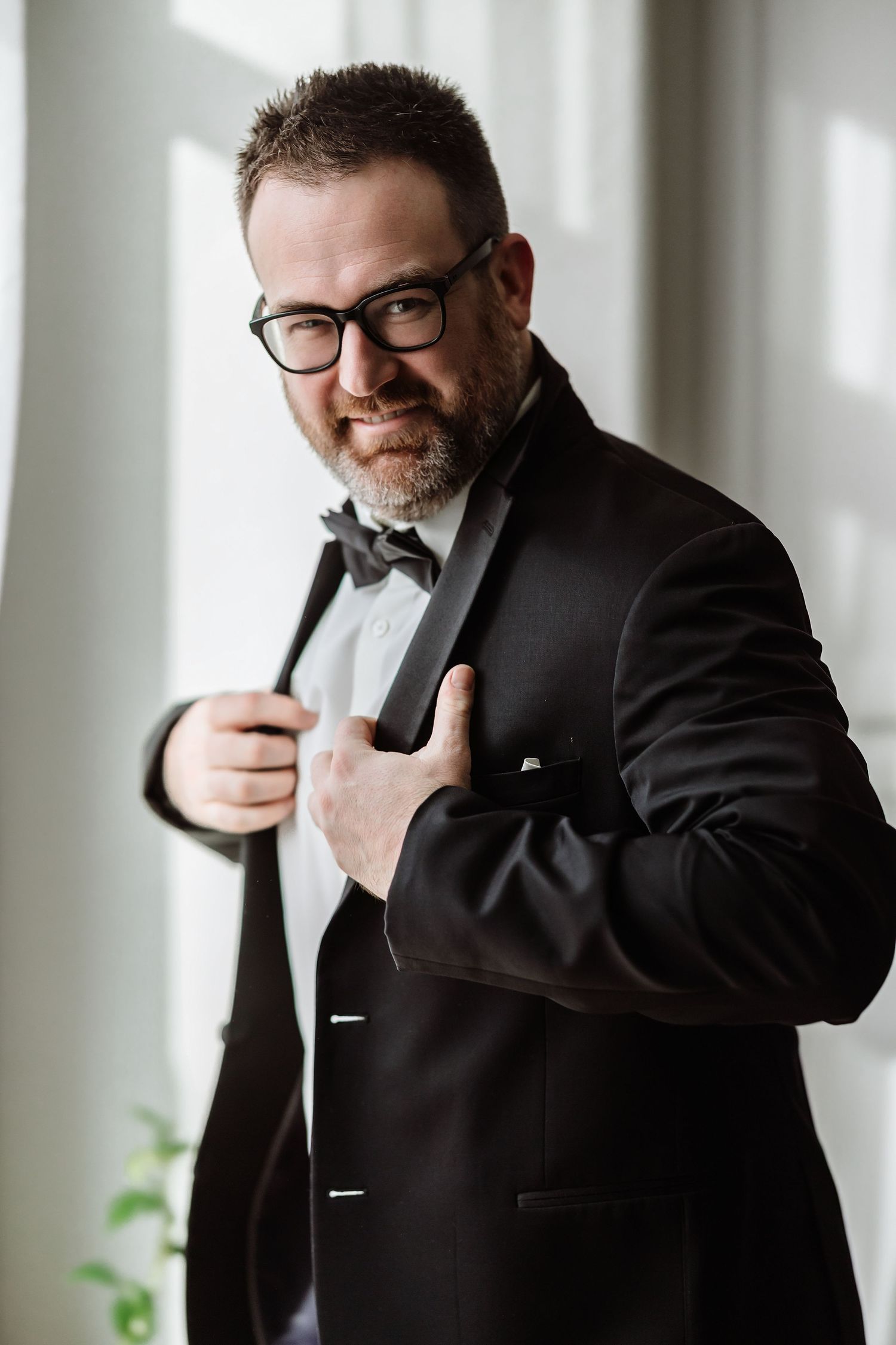 Smiling groom in black tuxedo adjusts his lapel while standing near a bright window with soft light.