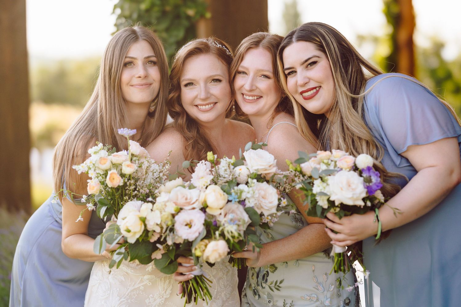 Bride and three bridesmaids in blue dresses smile together holding white and lavender bouquets.