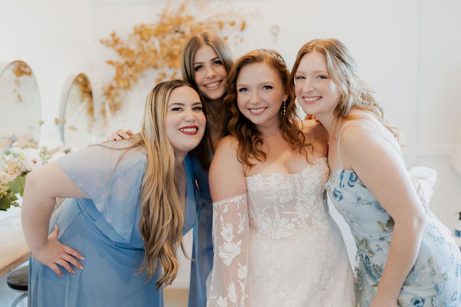Bride in lace gown poses joyfully with three bridesmaids in blue dresses indoors.