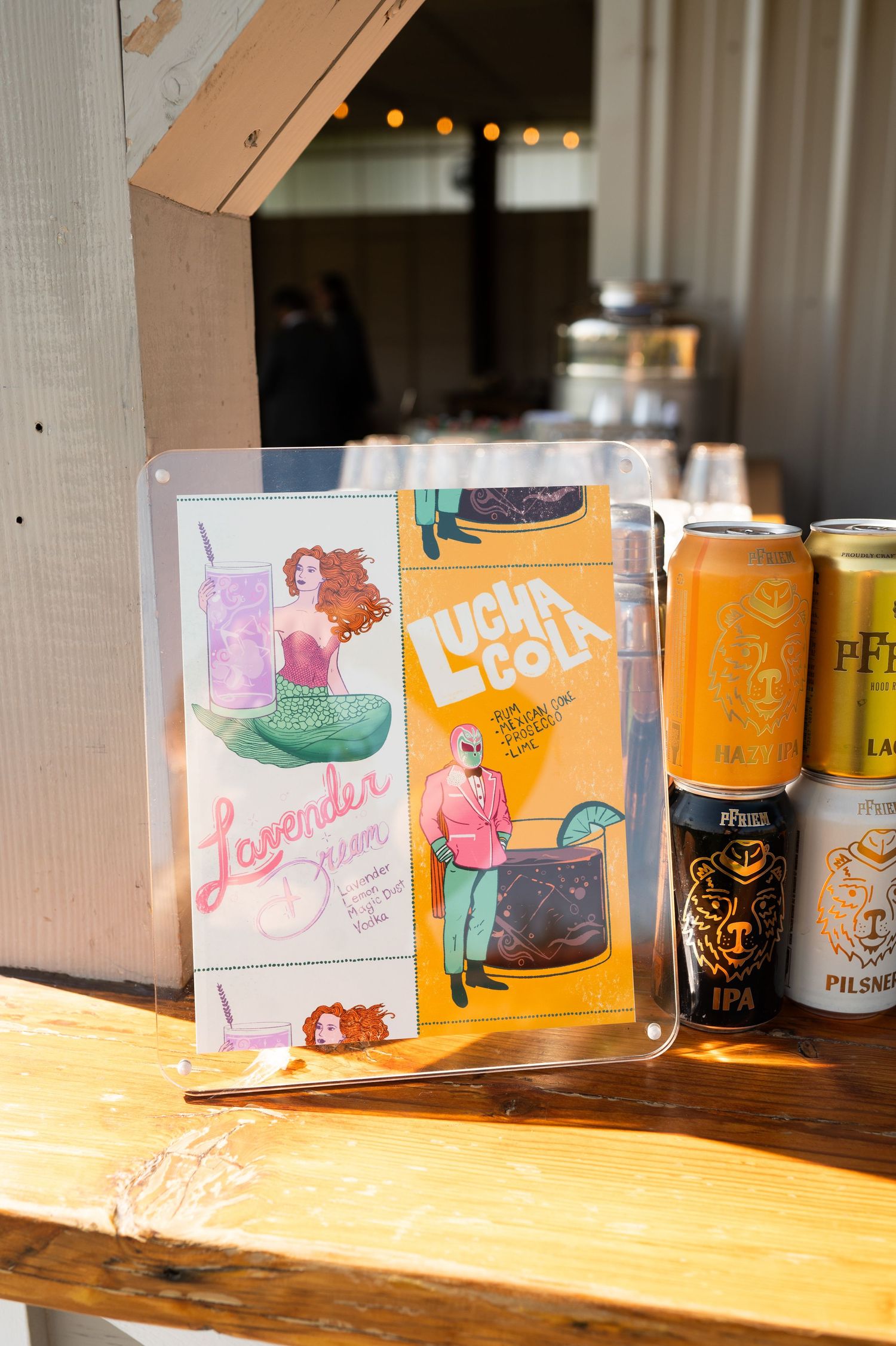 Colorful cocktail menu sign featuring mermaid artwork beside canned craft beers on a rustic wooden bar.