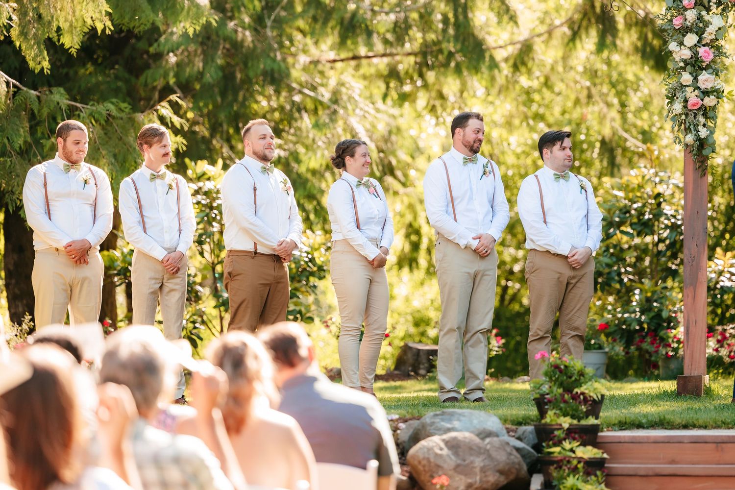 Six groomsmen in white shirts, tan pants, and suspenders stand at outdoor wedding ceremony.