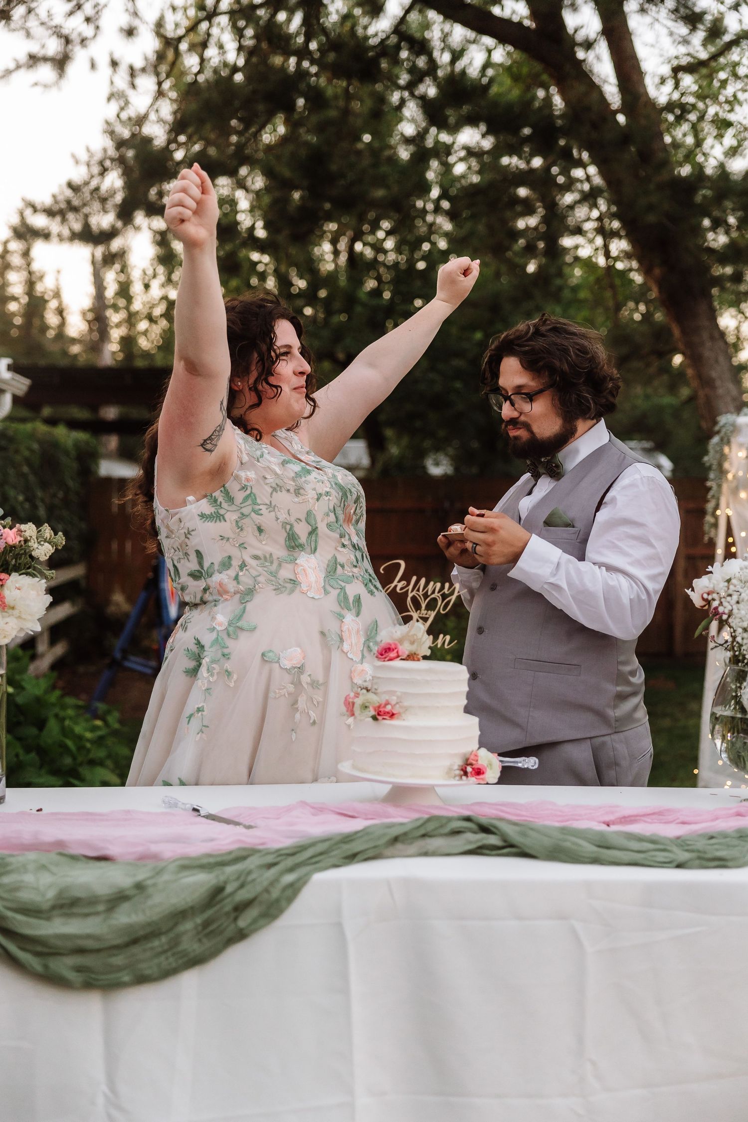 Bride raises arms in celebration while groom smiles during outdoor wedding cake cutting ceremony.