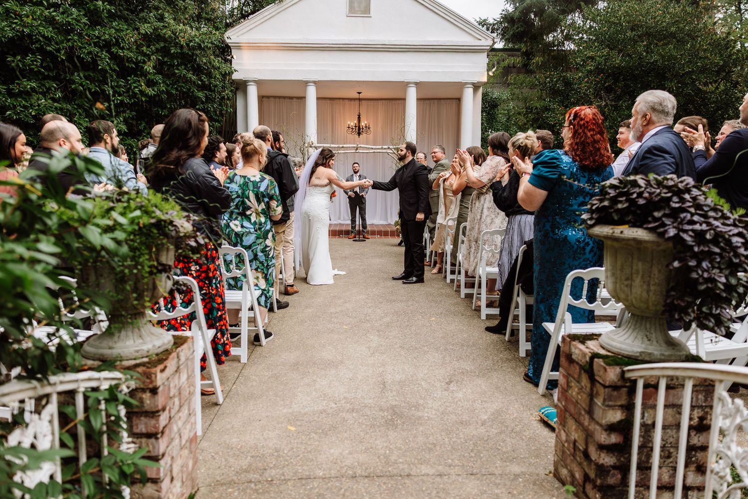 Outdoor wedding ceremony with guests standing along aisle leading to white columned chapel surrounded by lush greenery.