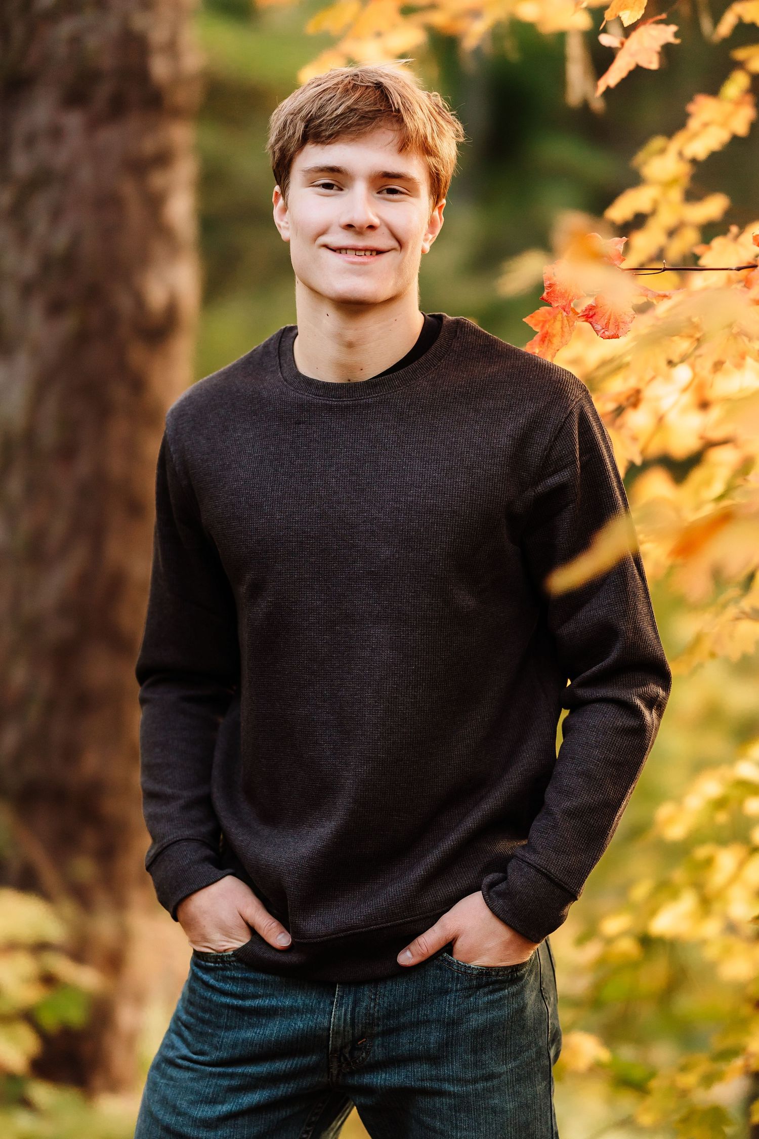 Young man in black sweatshirt and jeans smiling outdoors among vibrant autumn foliage in a forest setting.