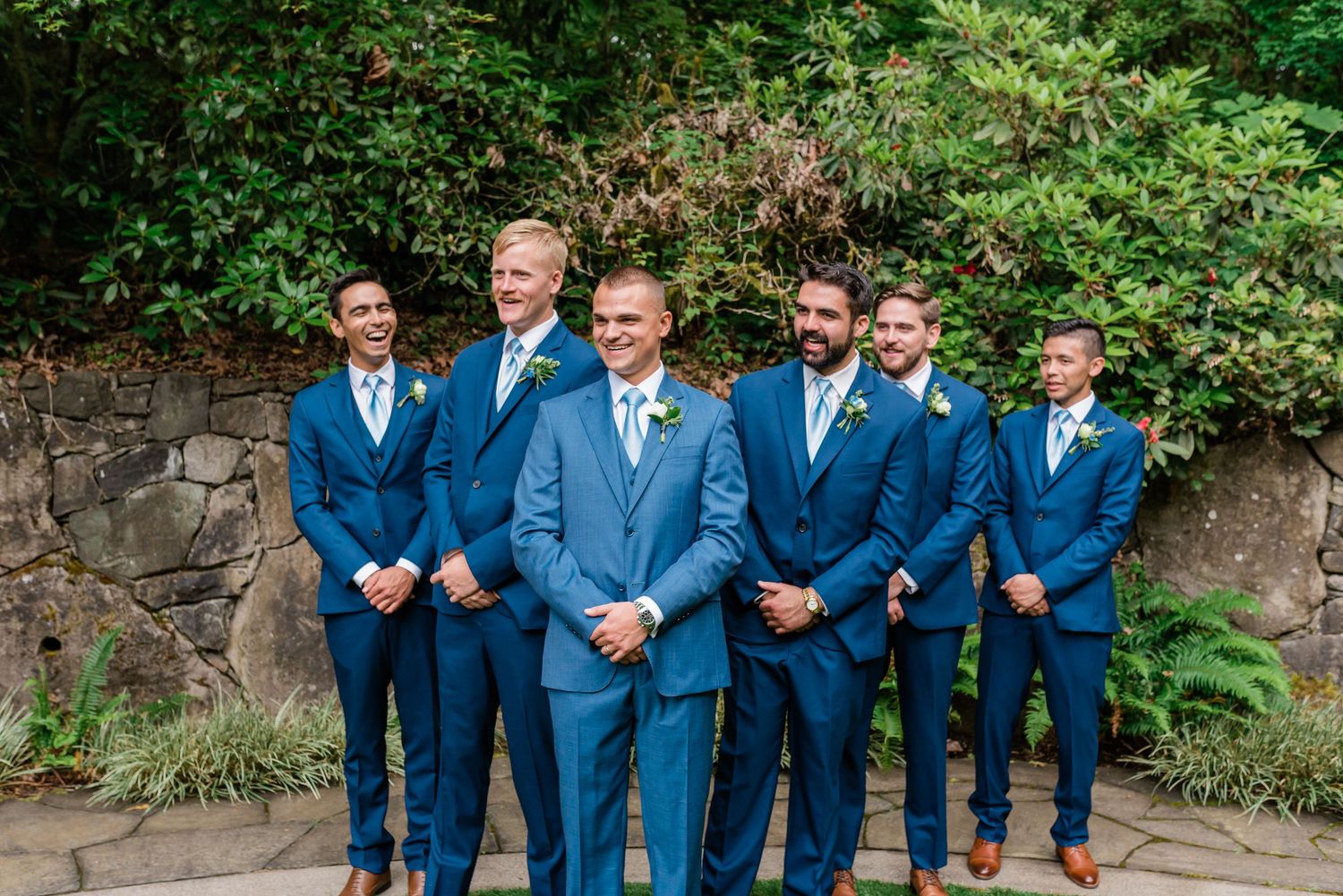 Groom with five groomsmen in matching navy blue suits posing before stone wall backdrop.
