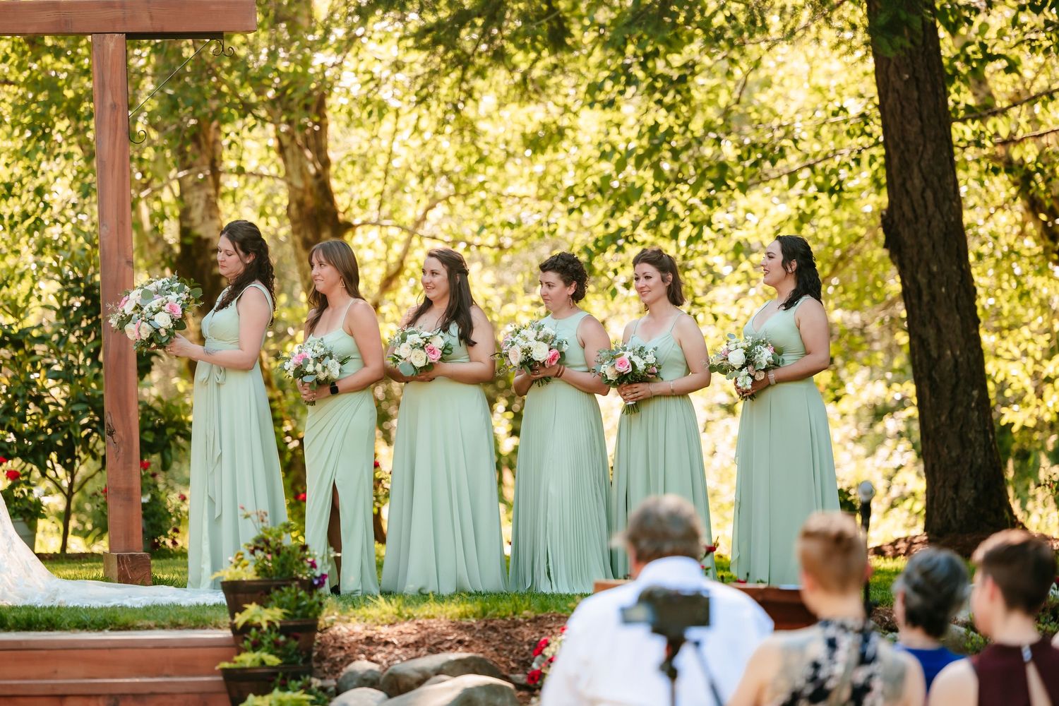 Five bridesmaids in sage green gowns holding bouquets stand beneath wooden arch at outdoor ceremony.
