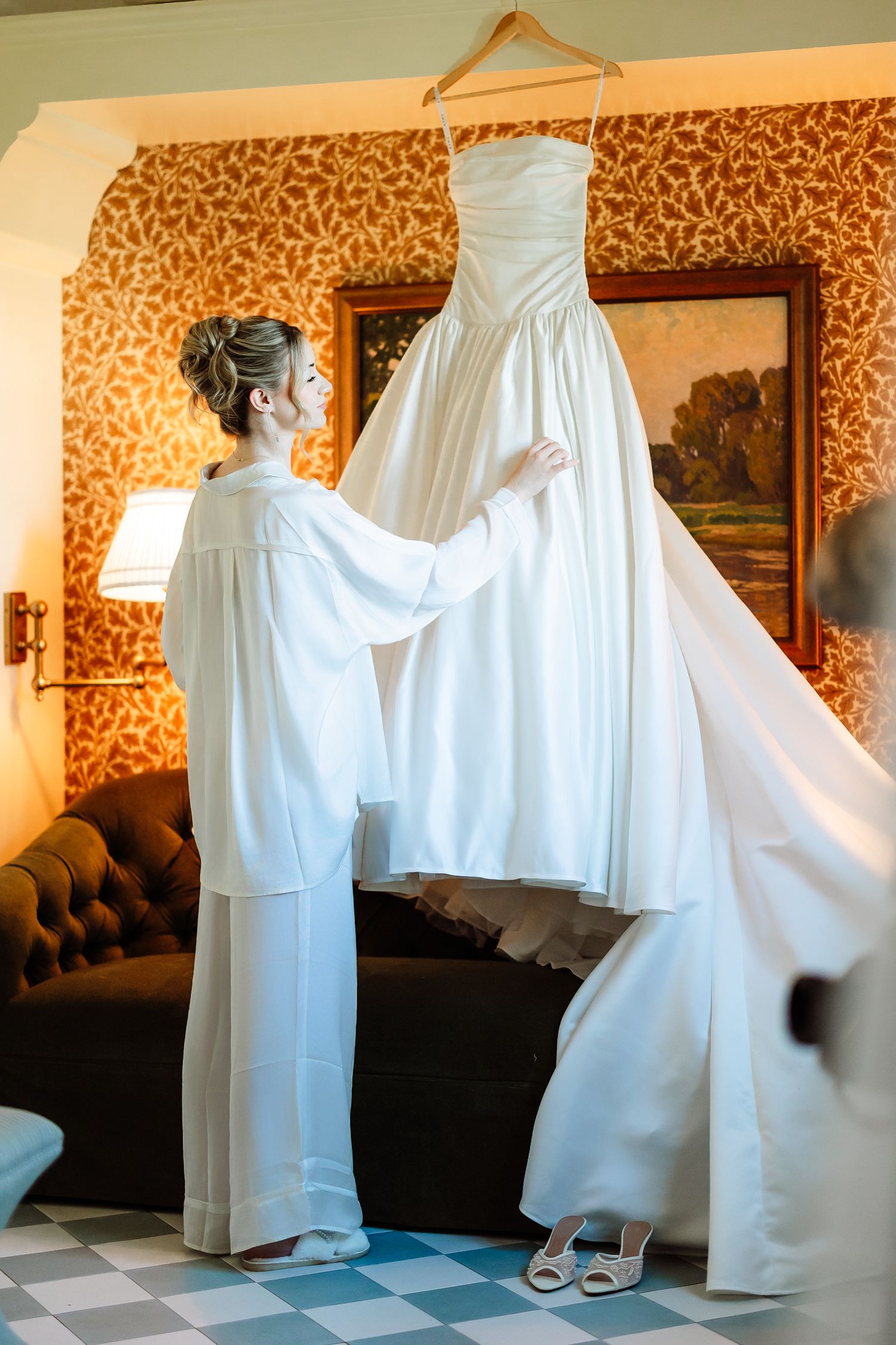 Bride in white robe admiring her strapless wedding gown hanging in a warmly decorated hotel room.