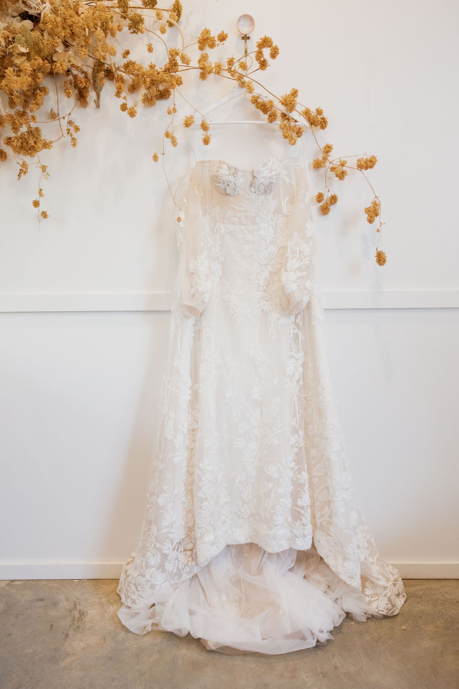 Elegant white lace wedding dress with long sleeves hanging against a white wall with dried floral arrangement.
