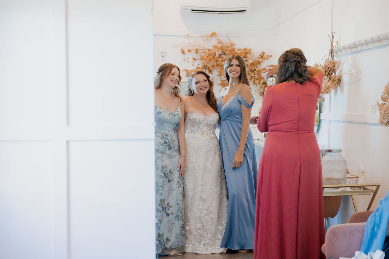 Bride in lace gown laughs with bridesmaids in blue dresses during wedding preparations.