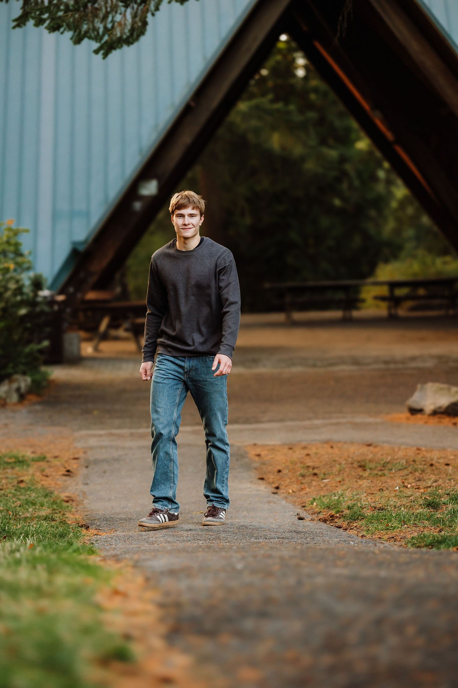 Young man in black sweater and jeans standing on a path in front of an A-frame building outdoors.
