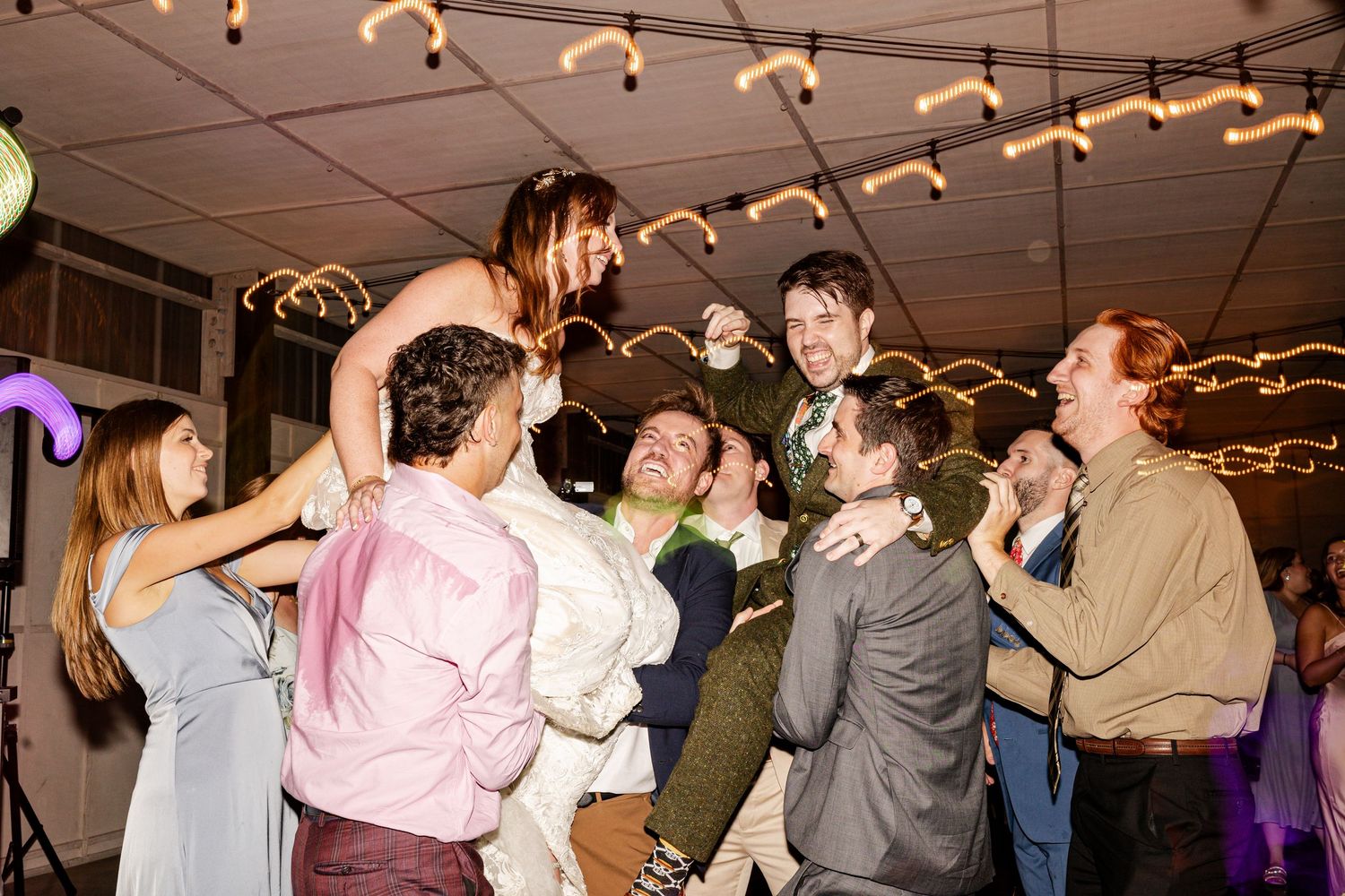 Joyful wedding reception guests dancing and celebrating together under warm string lights.
