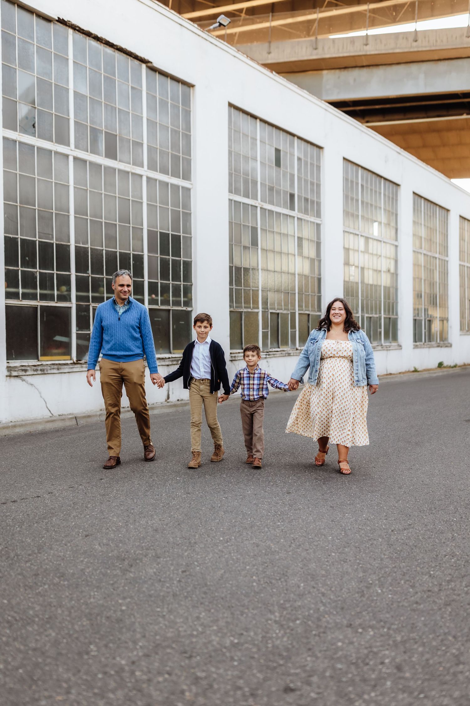 A happy family of four holds hands walking outside a white industrial building with large windows.