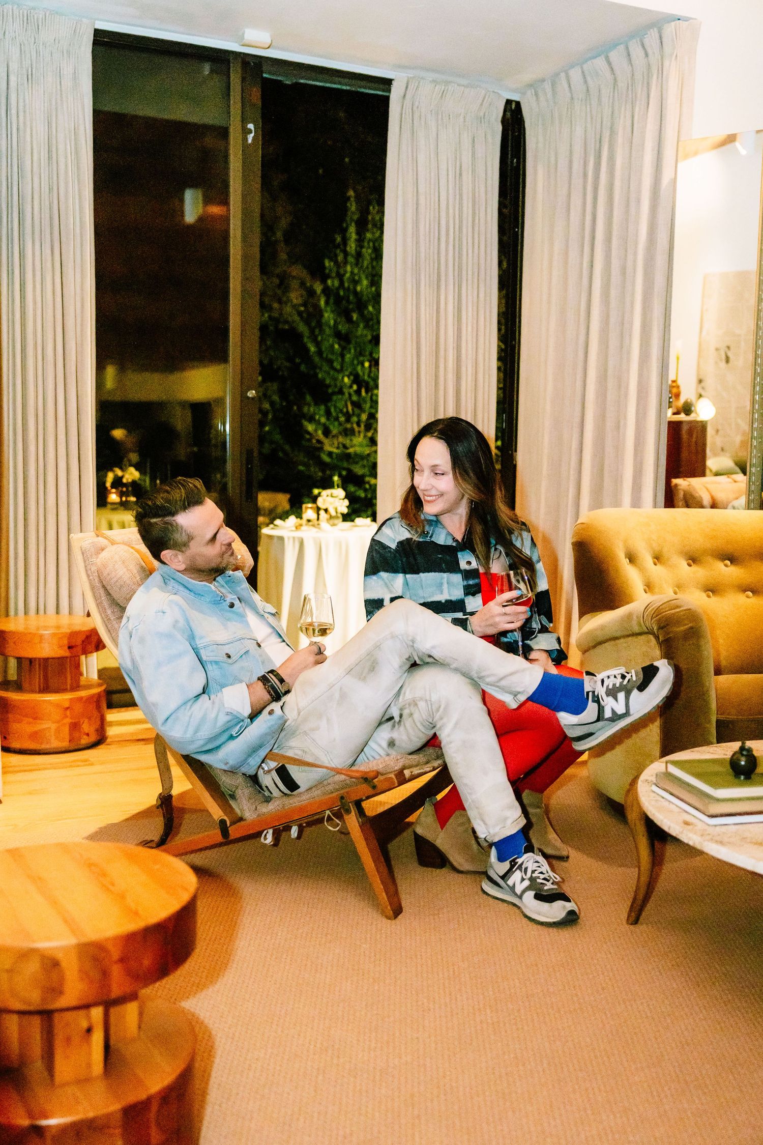 A man and woman sit in mid-century modern chairs, relaxing and chatting over drinks in a warmly lit living room.