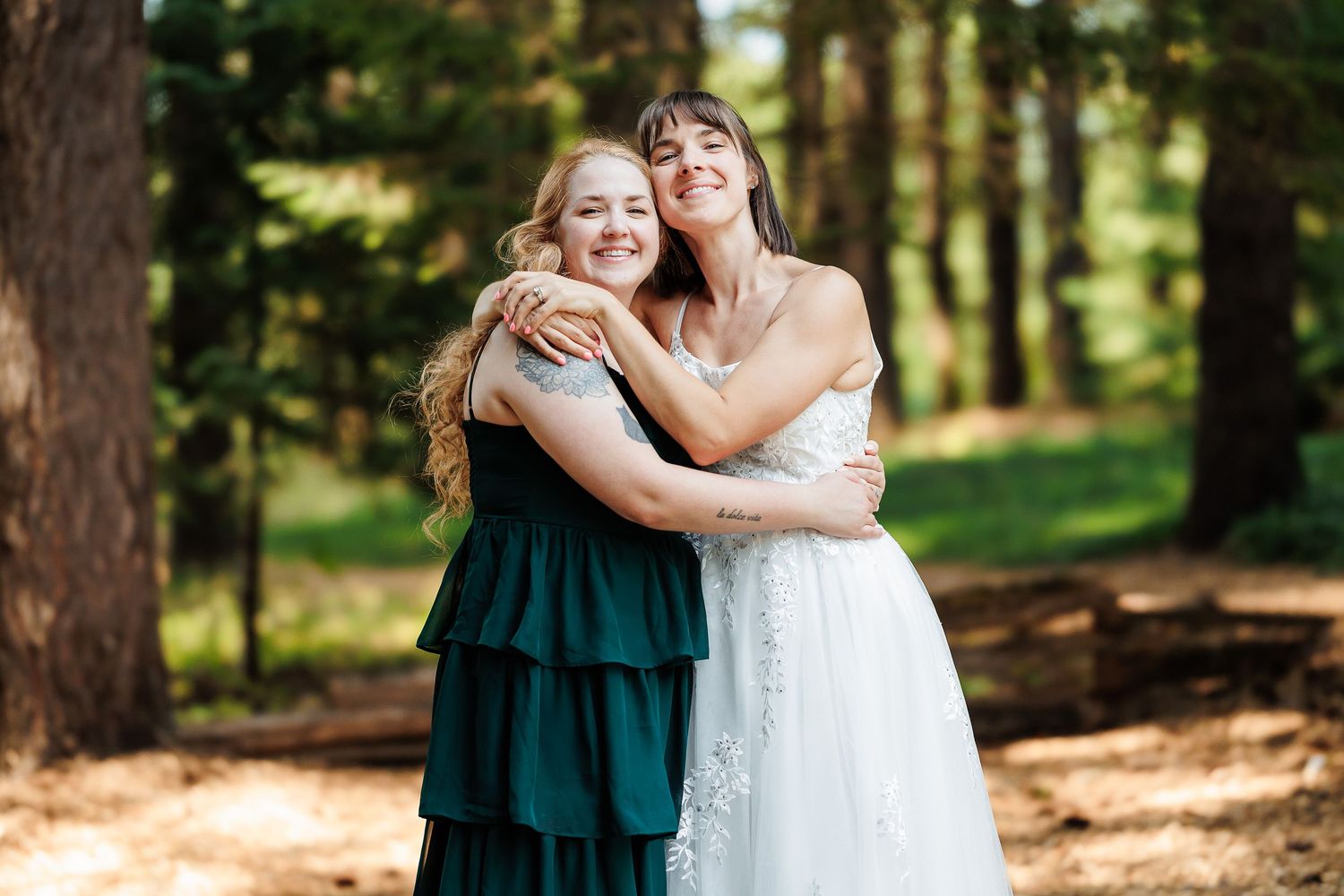 Bride in white lace gown hugs bridesmaid in emerald green dress, smiling warmly in a sunlit forest.