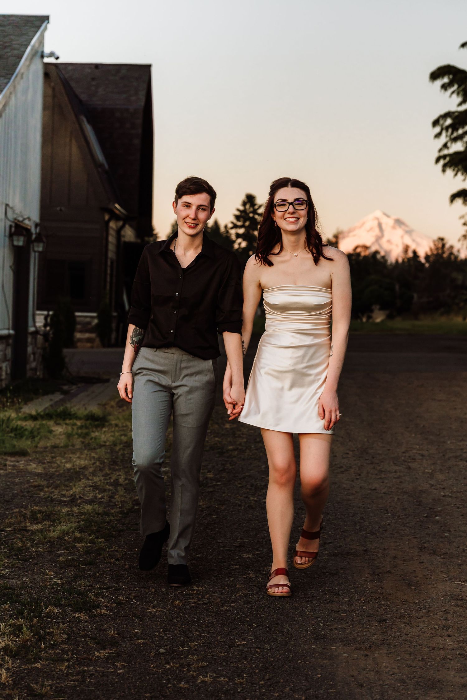 Couple walks hand in hand at sunset near a barn with a mountain visible in background.