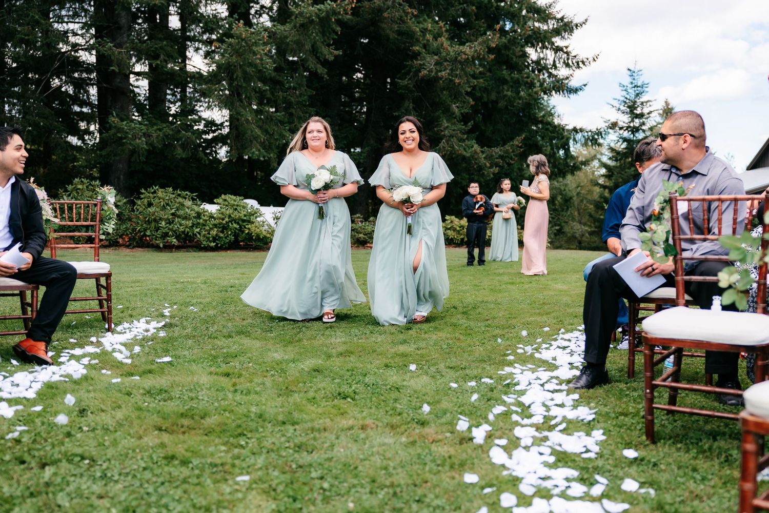 Two bridesmaids in sage green gowns walk down petal-lined aisle at outdoor garden wedding ceremony.