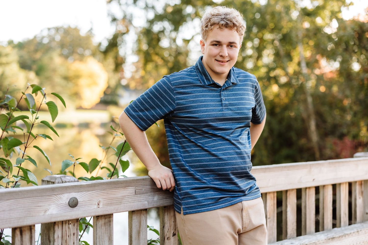 Smiling young man in blue striped polo leaning on wooden bridge railing outdoors in autumn setting.