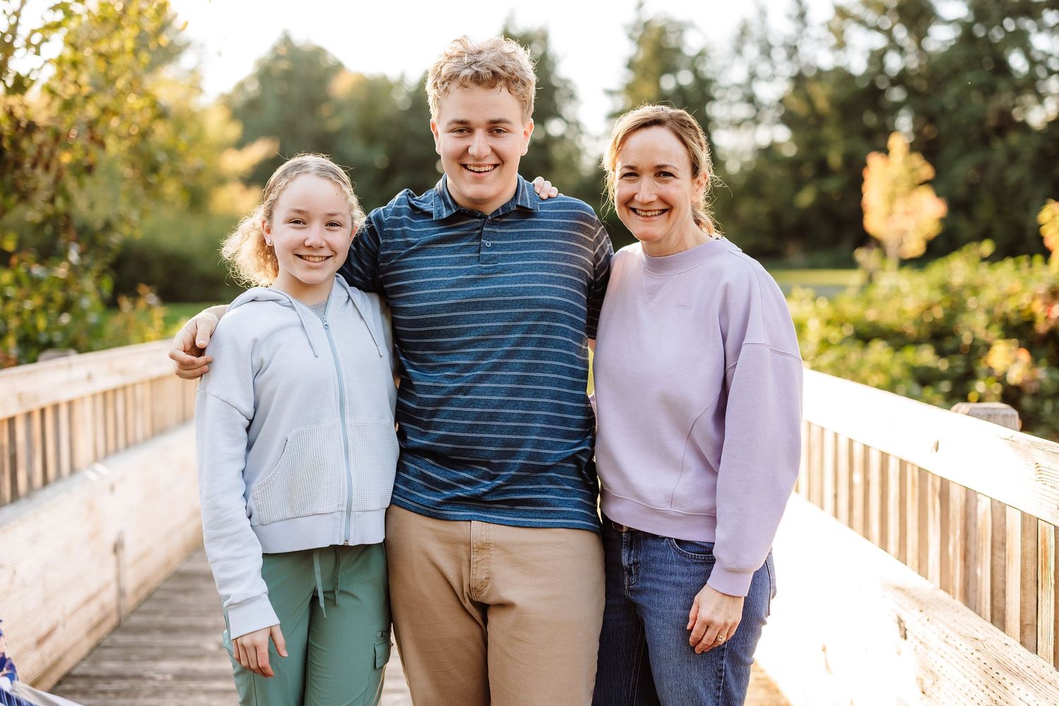 A family of three smiling on a wooden bridge in a sunny park, with a young girl, teenage boy, and woman.