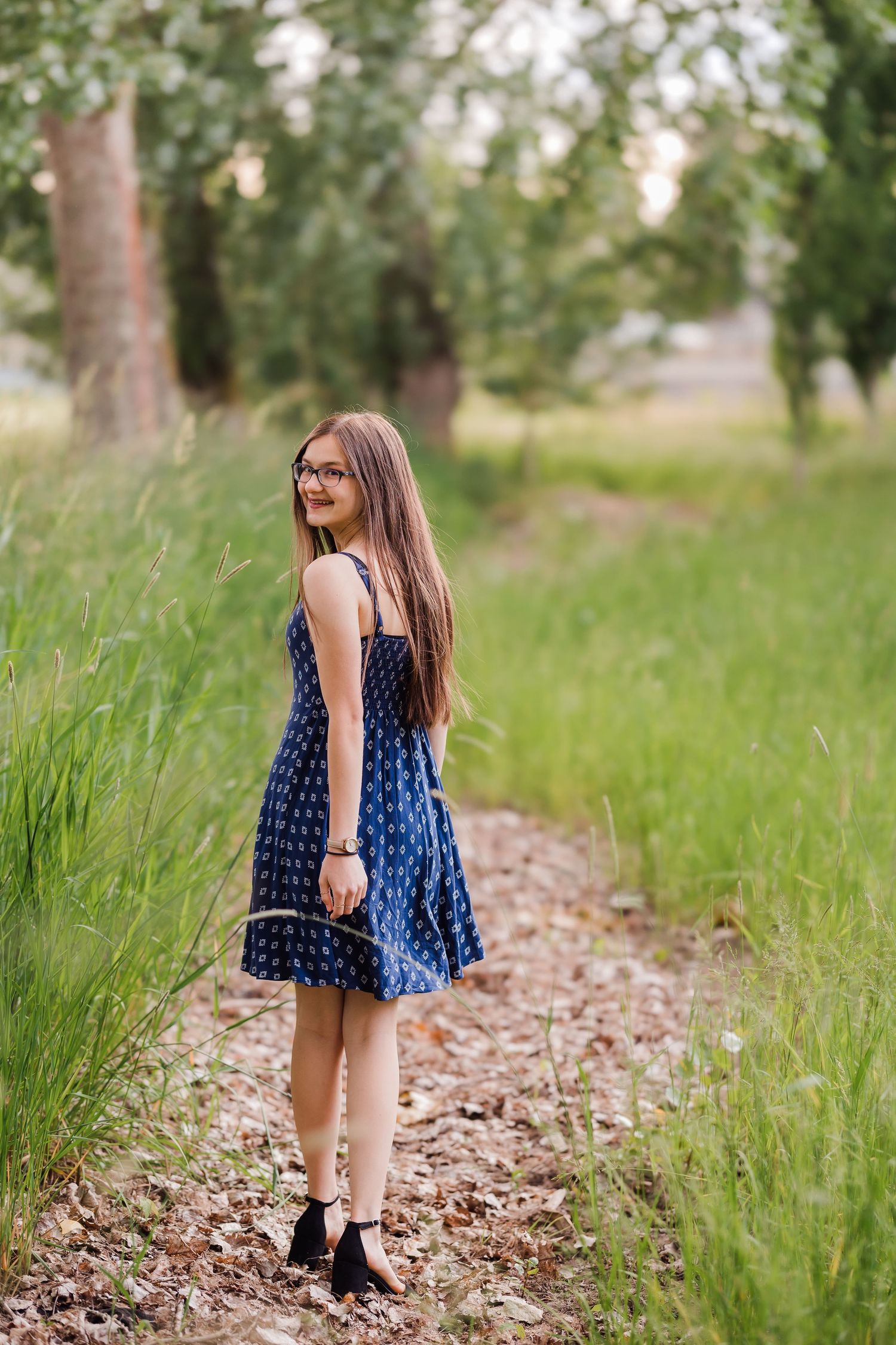 Young girl in blue sundress and glasses smiling over shoulder on nature path surrounded by green trees.
