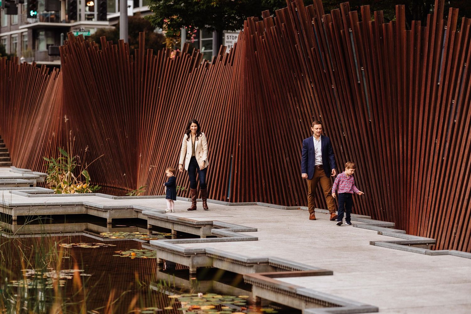 Family strolling through a modern urban plaza with tall rust-colored steel sculptures beside a reflecting pool.