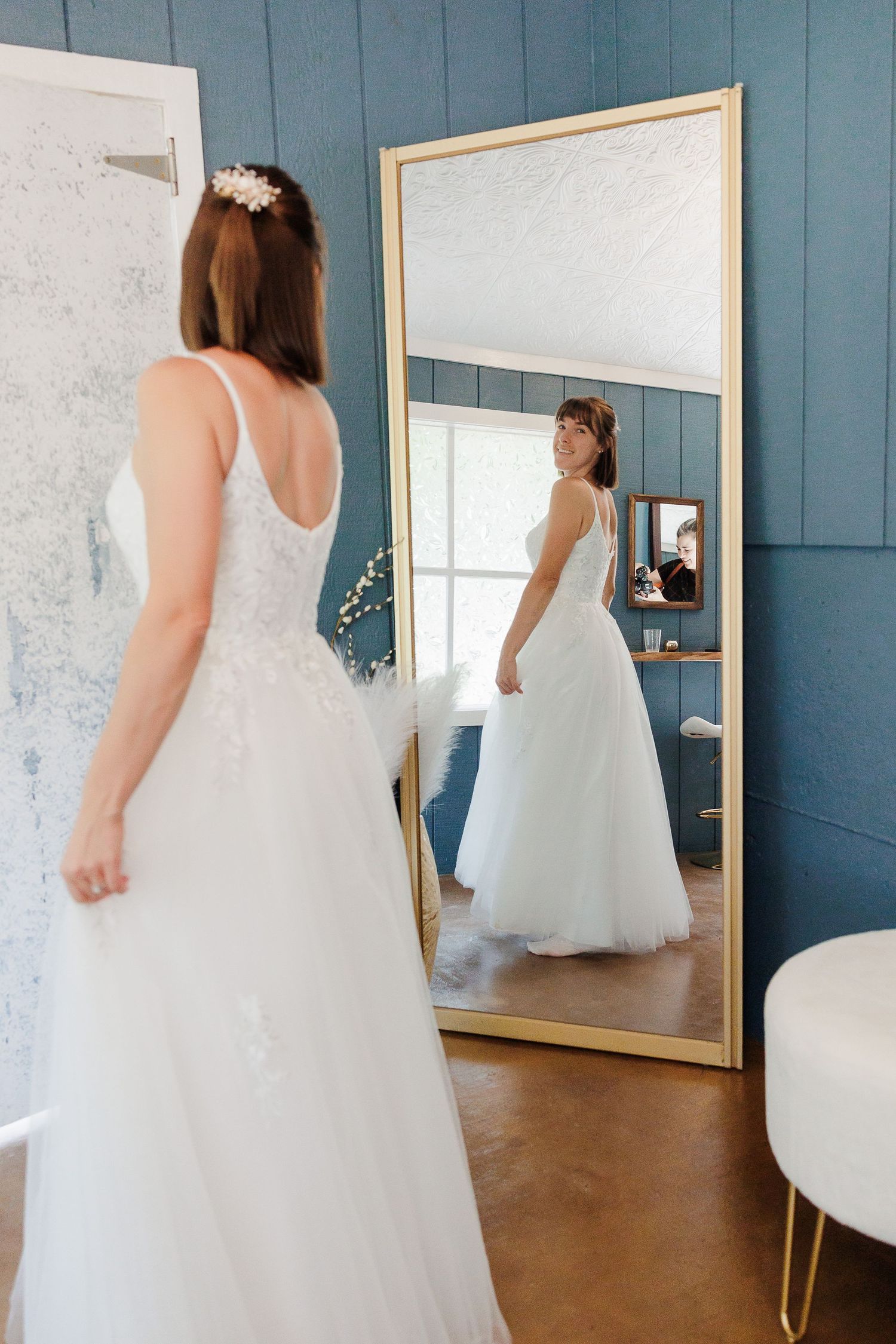 Bride in a white lace wedding gown admiring her reflection in a large gold-framed mirror.