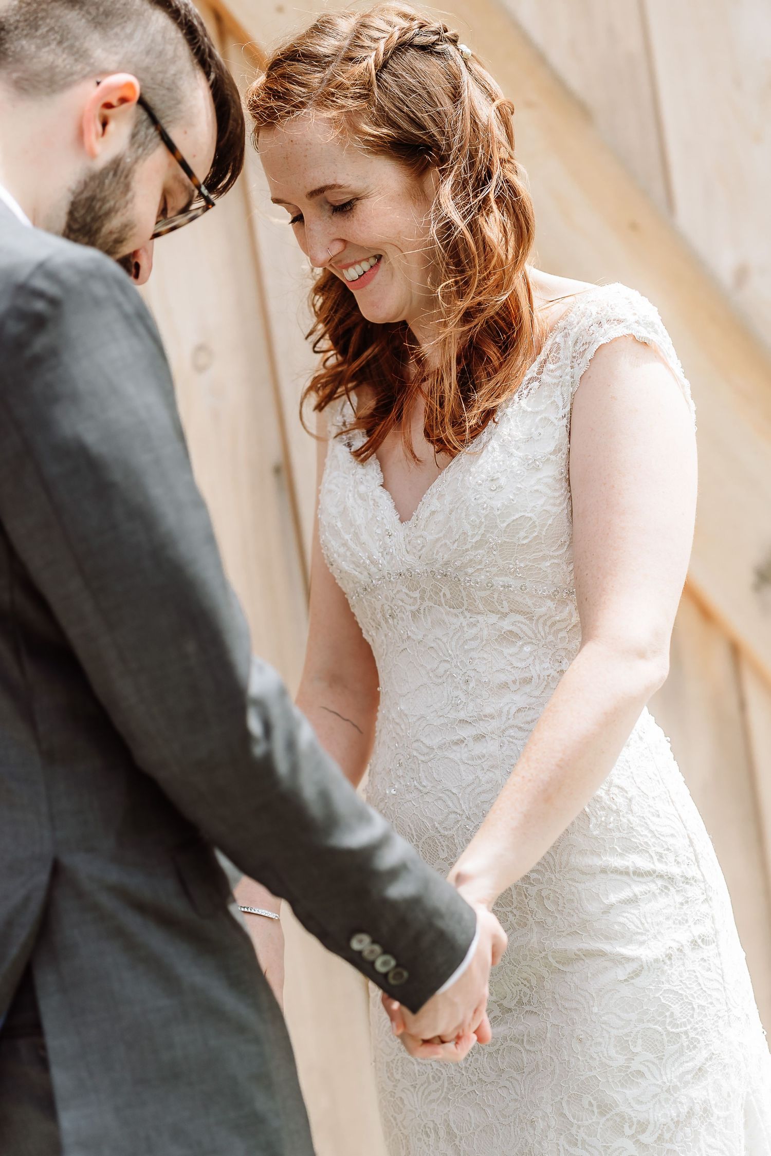 Smiling bride in lace cap-sleeve gown holds hands with groom near wooden barn wall.