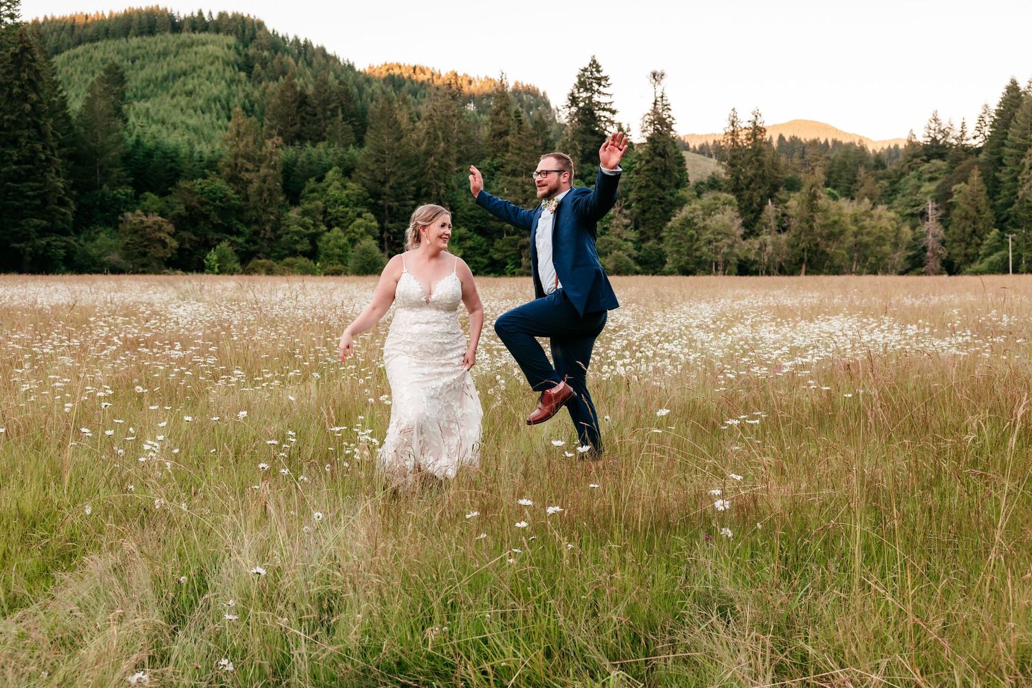 Joyful bride and groom running and jumping through a meadow of wildflowers with forested hills in background.