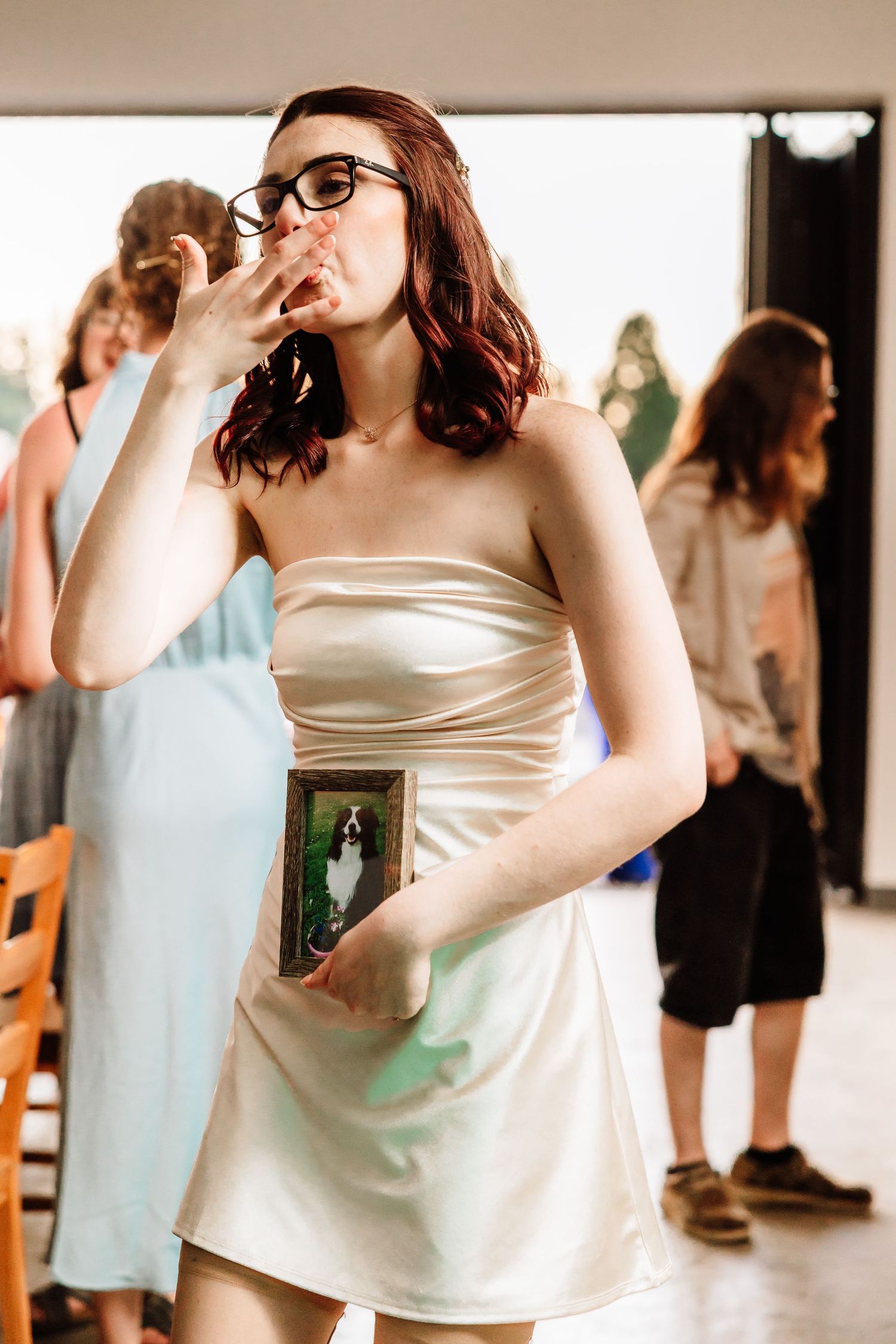 Bride in white strapless dress holding a framed pet photo at wedding reception.
