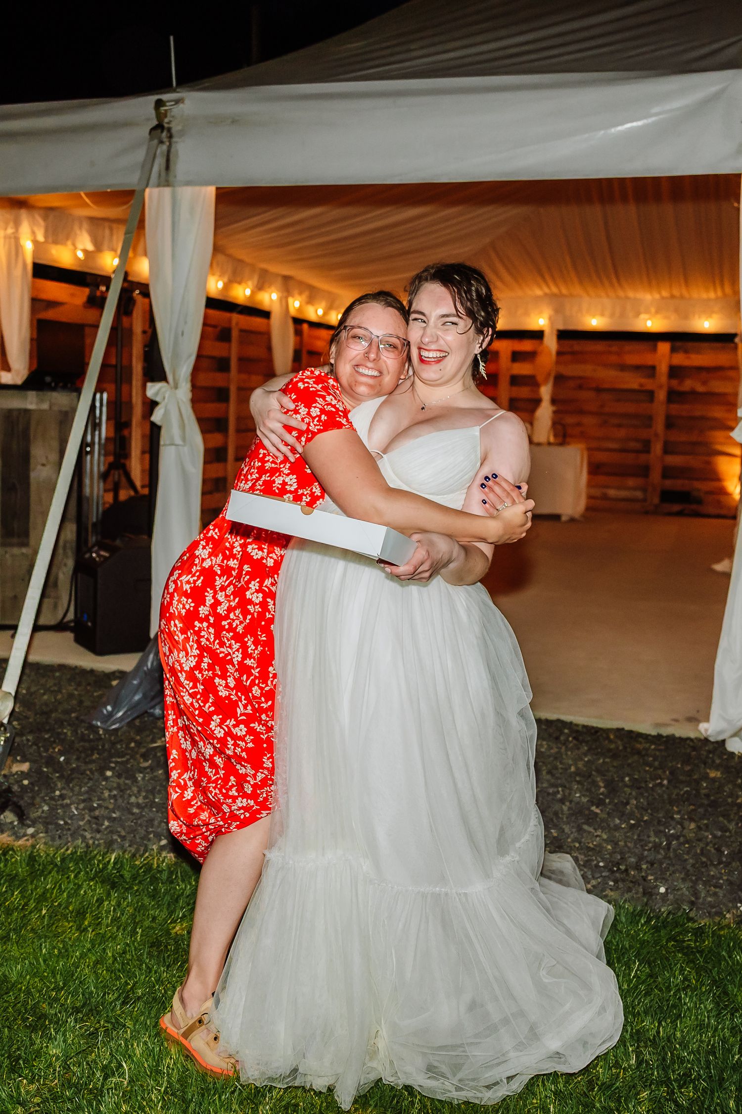 Bride and female guest sharing a warm hug outside a wedding reception tent at night.