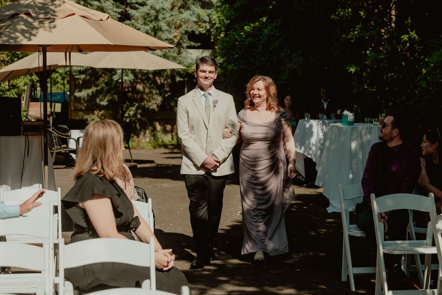 A young man in a suit escorts a woman in a silver gown down the aisle at an outdoor wedding ceremony.