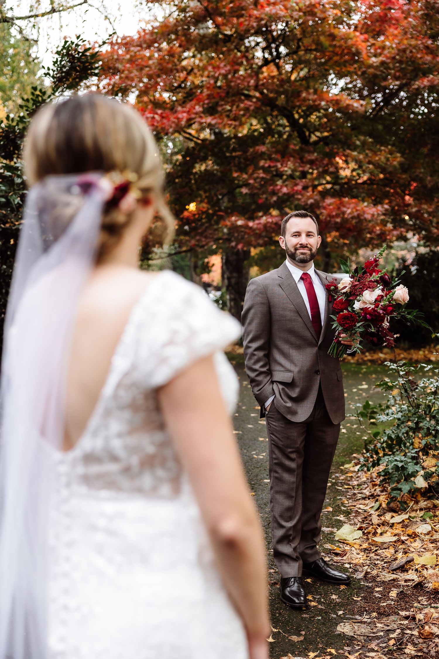 Groom smiles at bride during autumn outdoor first look with red foliage.