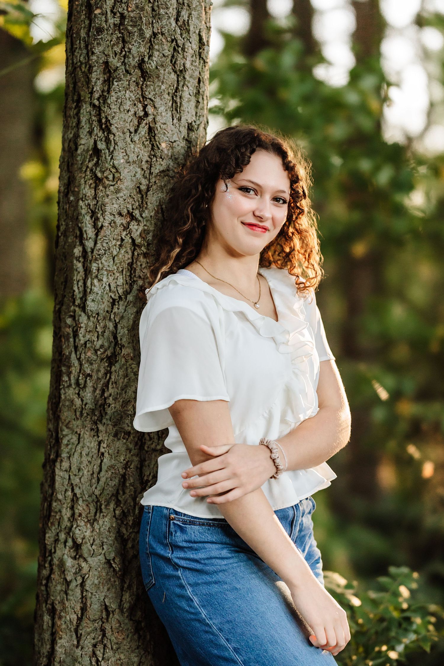 Young woman with curly hair in white blouse and jeans leaning against a tree in a sunlit forest.
