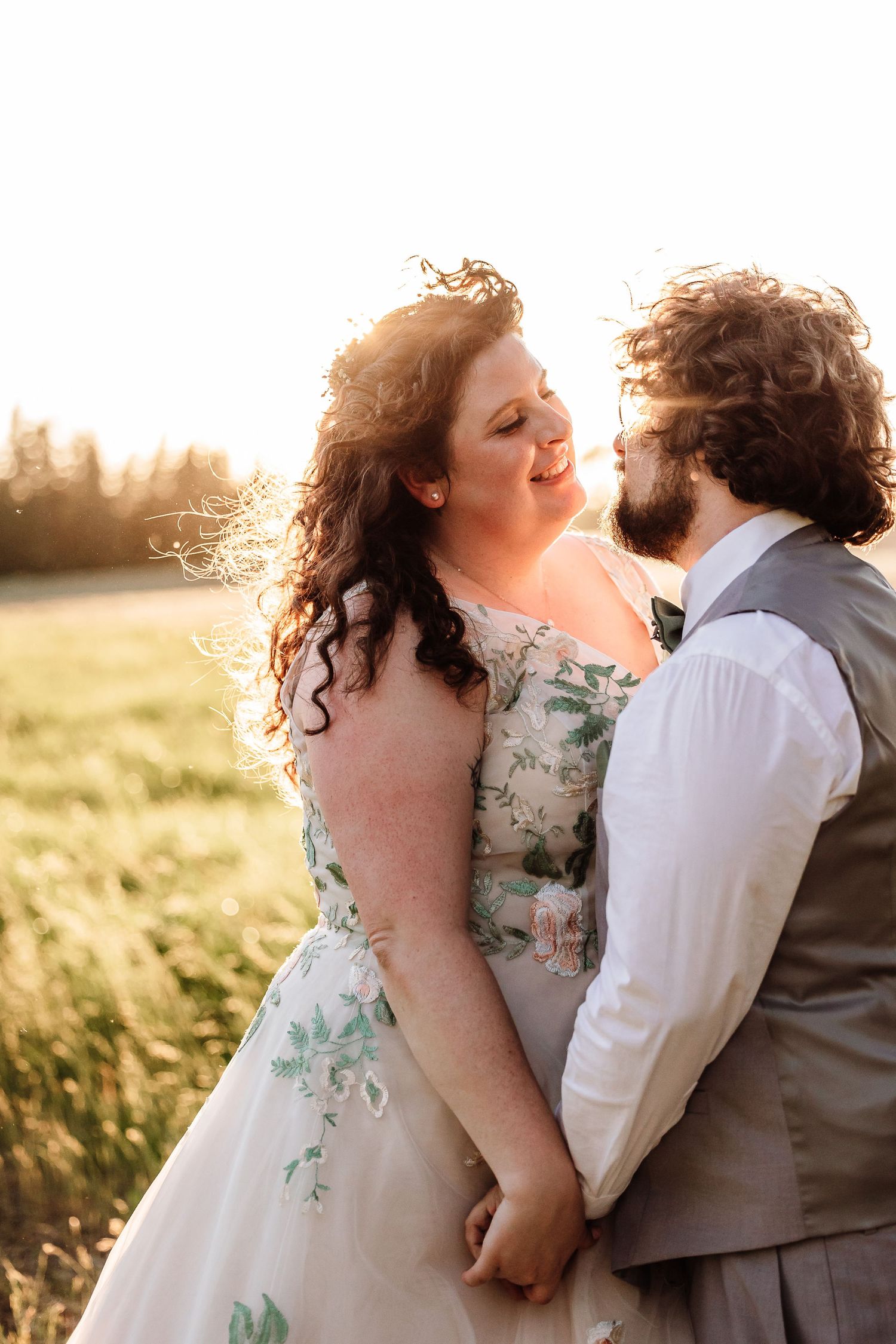 A bride and groom holding hands and gazing at each other in a sunlit meadow during golden hour.