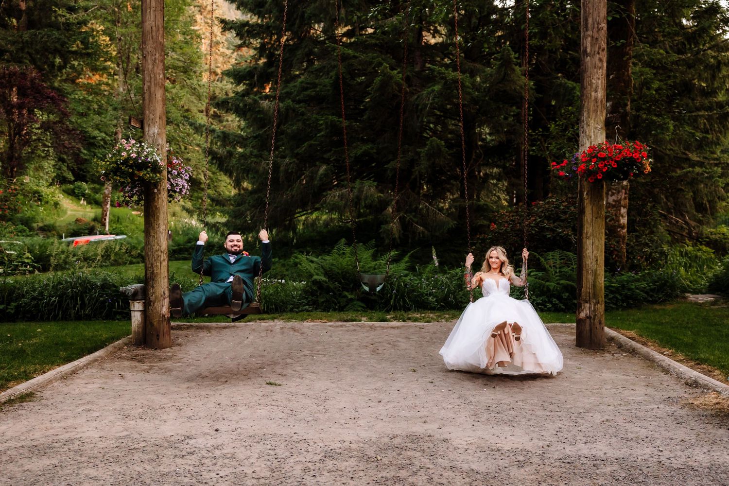 Bride and groom joyfully swinging on large outdoor swings surrounded by tall trees and flowers.