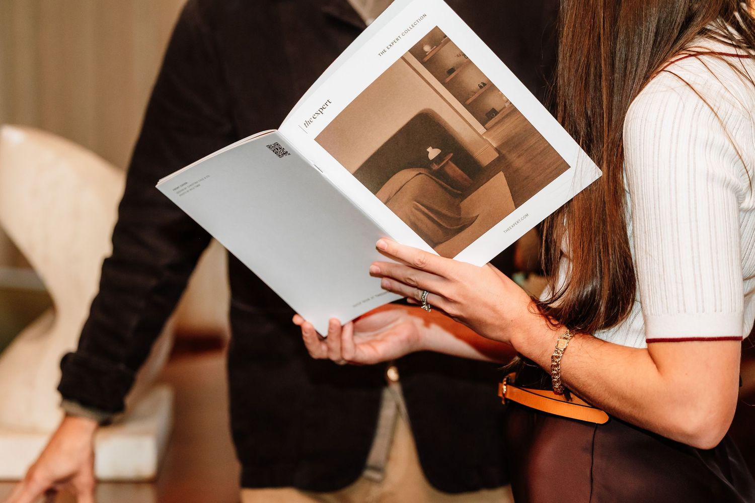 A woman holds and reads an open interior design catalog or brochure at an event.