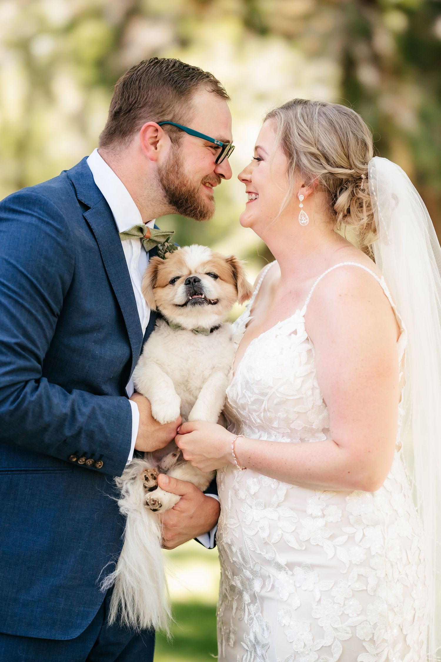 Smiling bride and groom holding a small fluffy dog between them while looking at each other outdoors.