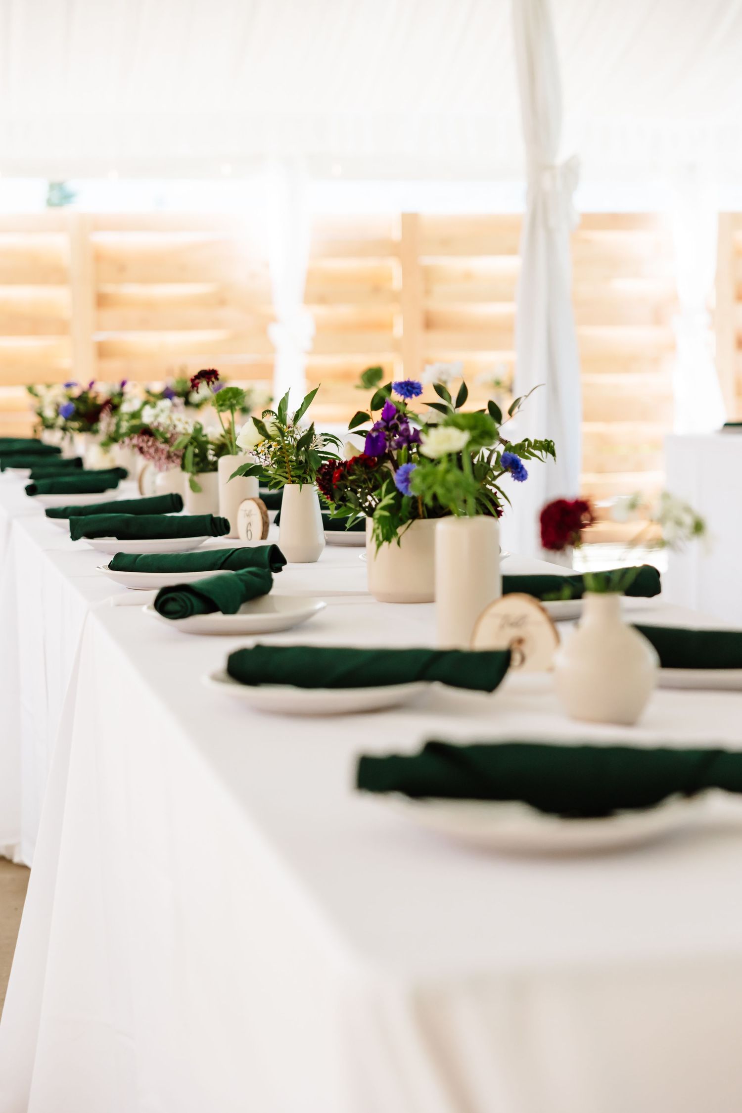 Wedding reception head table with white linens, green rolled napkins, and small floral arrangements in cream vases.