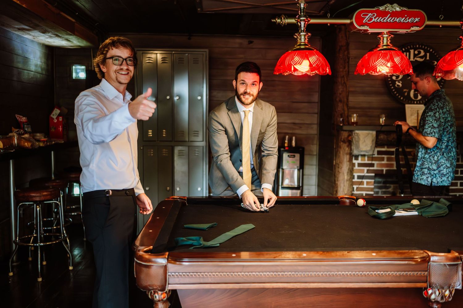 Two men smiling near a pool table under red Budweiser hanging lights.