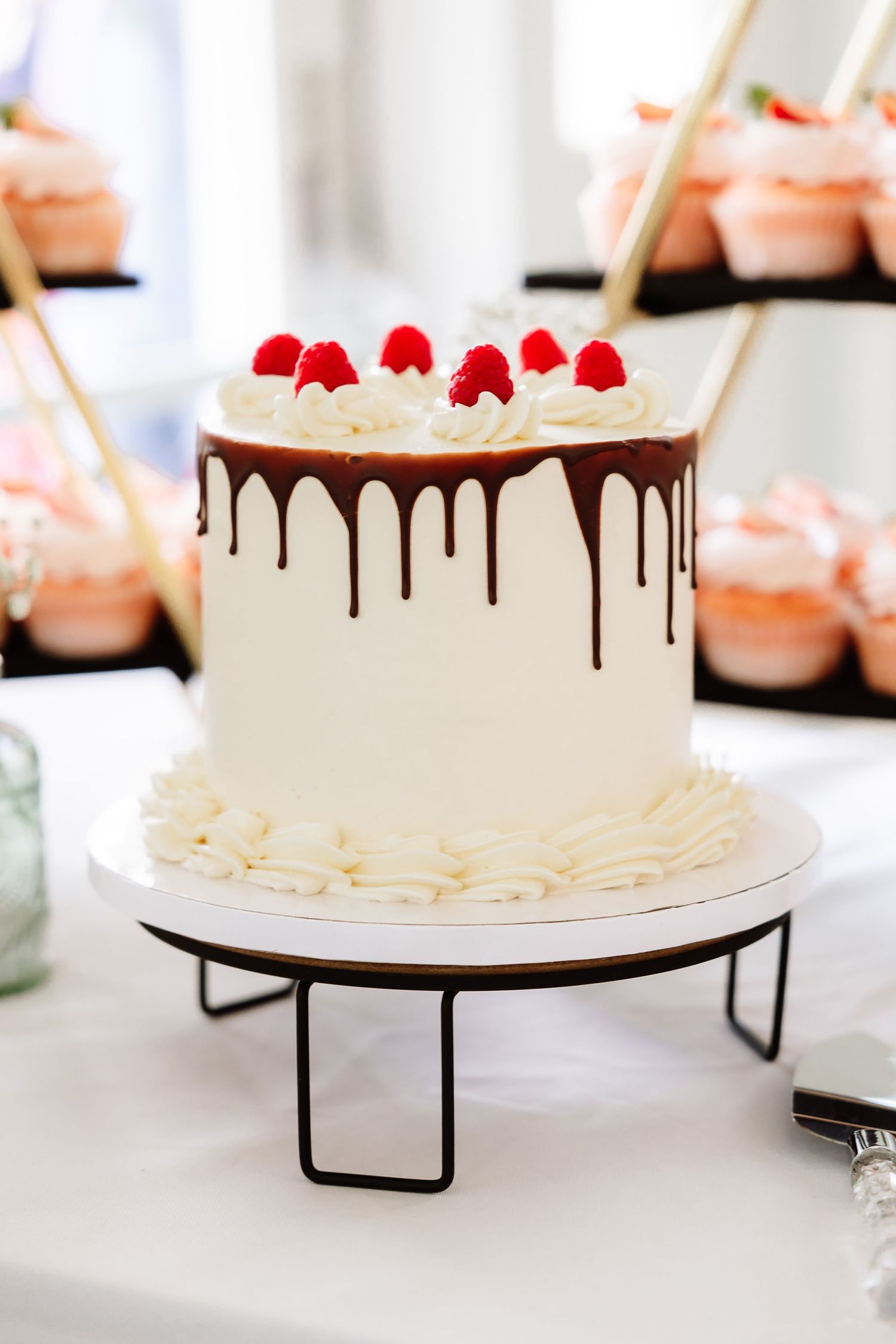 White wedding cake with chocolate drip, raspberry topping, and pink cupcakes in background.