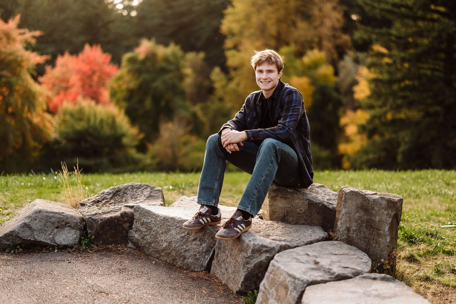 A young man sits smiling on stone steps in a park with colorful autumn foliage in the background.