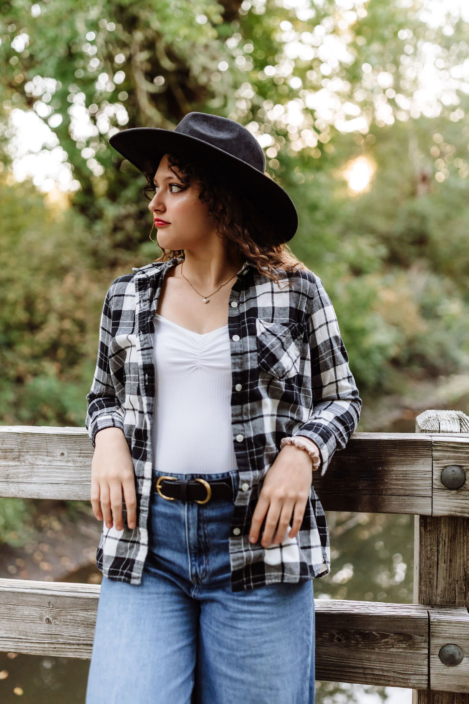 Woman in plaid shirt, white top, jeans, and wide-brim hat posing on a wooden bridge surrounded by greenery.